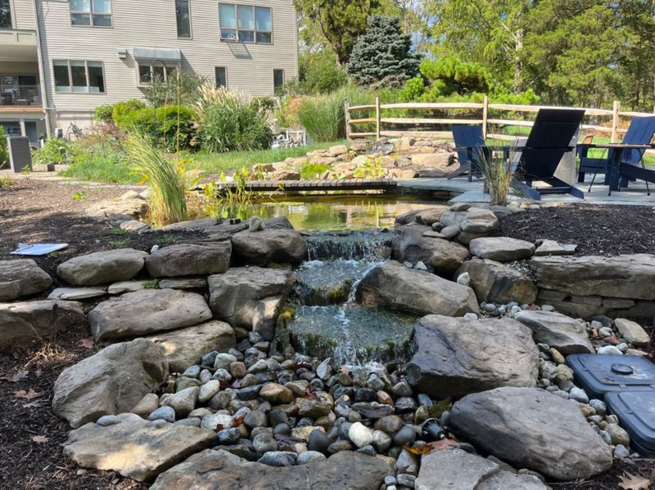Backyard pond and waterfall with patio seating and a wooden bridge by AquaReale Philadelphia
