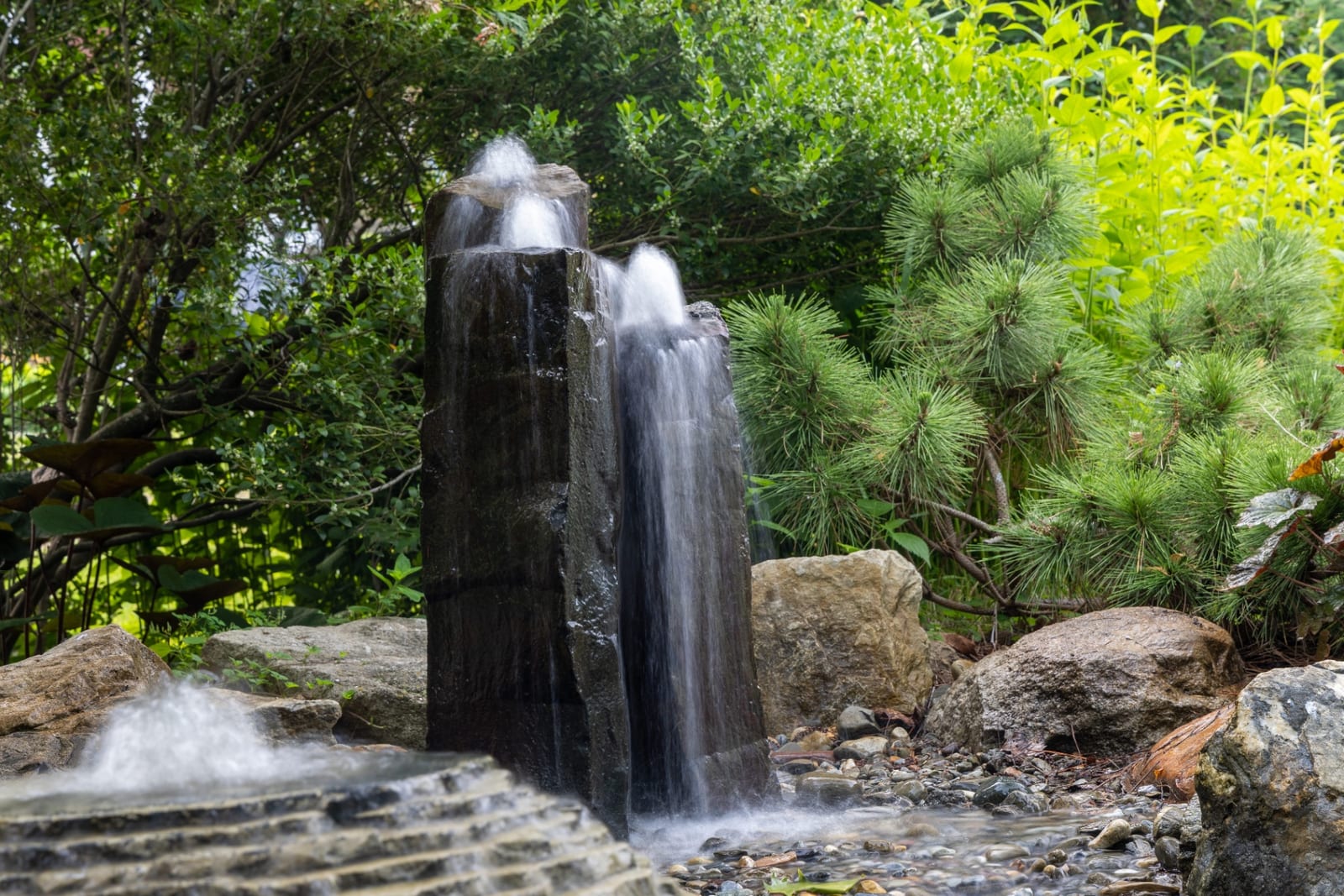 Basalt column fountain with cascading water among pines by AquaReale Philadelphia