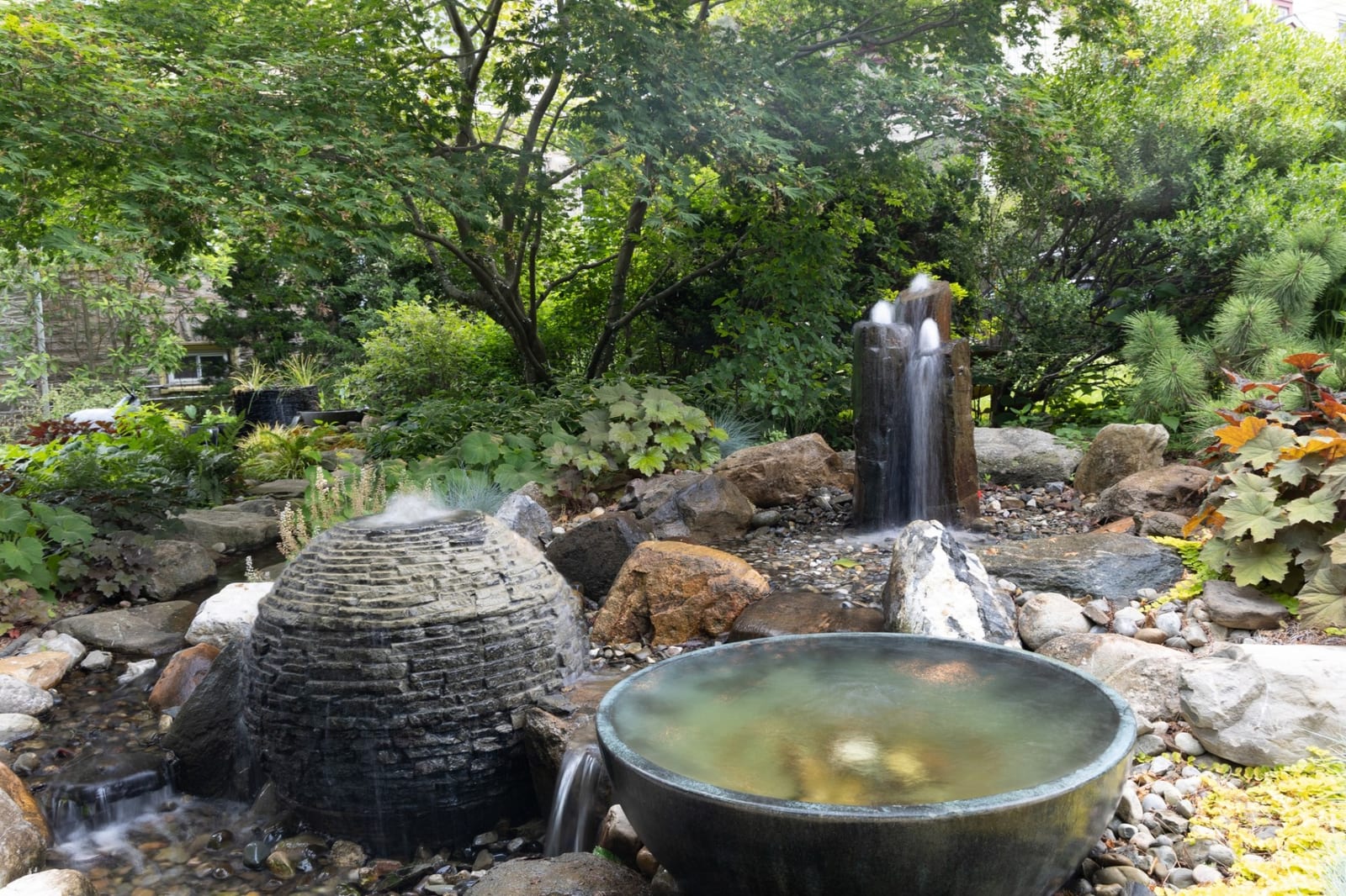 Cluster of decorative bubbling urns, basalt column fountain and ceramic bowl water features in lush garden setting by AquaReale