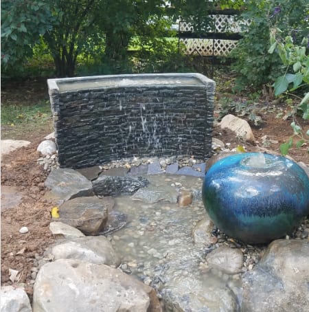 A stacked stone spillway wall and a glazed blue sphere fountain in a backyard pebble bed — a low-maintenance decorative fountainscape installed by AquaReale near Philadelphia