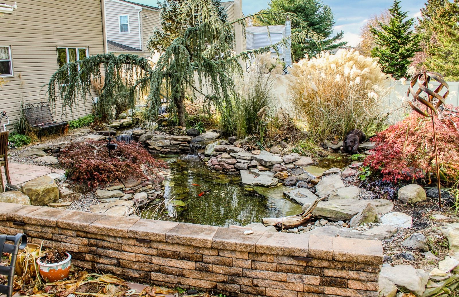 Backyard koi pond with stacked stone retaining wall and weeping tree by AquaReale Philadelphia