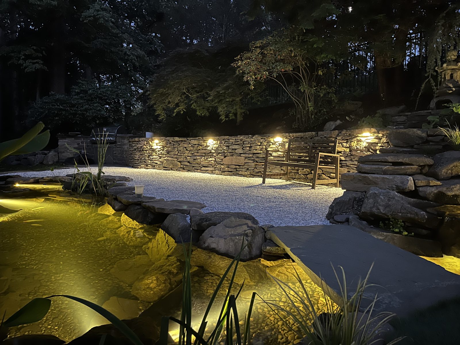 Backyard pond at dusk with warm landscape lighting along the path by AquaReale Philadelphia